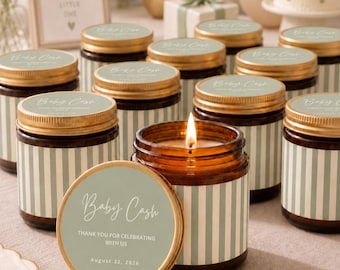 May include: A collection of amber glass candle jars with gold lids, each featuring a sage green and white striped label. The labels display the text Baby Cash, Thank You For Celebrating With Us, and the date August 22, 2026. One candle is lit.