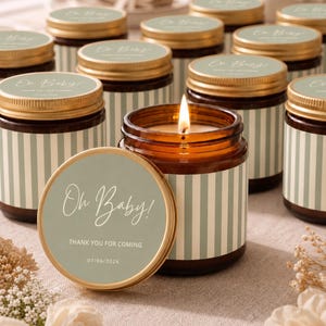 May include: A collection of amber glass candles with gold lids, each featuring a sage green label with white vertical stripes. The labels display the text Oh Baby! THANK YOU FOR COMING 07/06/2026. One candle is lit, showing a bright flame.