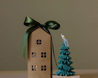 May include: A green candle shaped like a Christmas tree with a small tag that says "vetha" is on a wooden base. A brown cardboard house with cut-out windows is wrapped with a green ribbon.