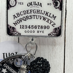 Puede incluir: Un portatarjetas rectangular con un dise&ntilde;o de tablero Ouija y un borde plateado brillante. El tablero presenta el alfabeto, n&uacute;meros y las palabras "S&iacute;", "No" y "Adi&oacute;s". Un dije de cuentas negras y otros dijes cuelgan debajo.