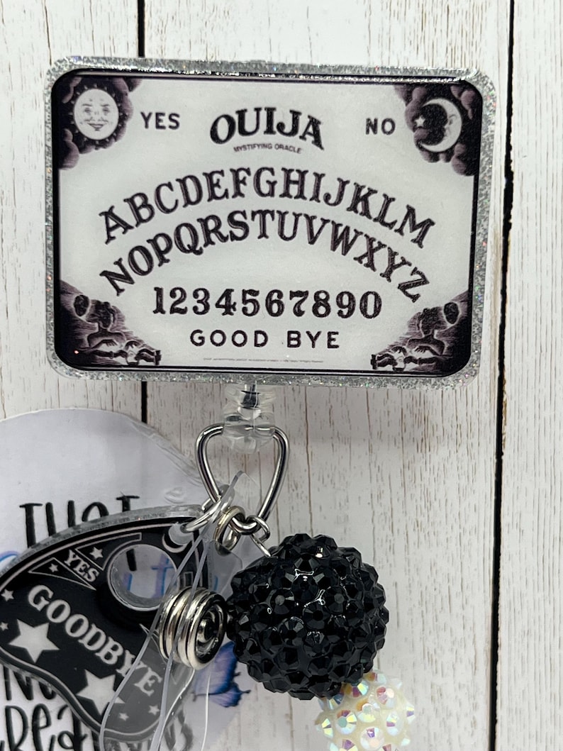Puede incluir: Un portatarjetas rectangular con un dise&ntilde;o de tablero Ouija y un borde plateado brillante. El tablero presenta el alfabeto, n&uacute;meros y las palabras "S&iacute;", "No" y "Adi&oacute;s". Un dije de cuentas negras y otros dijes cuelgan debajo.