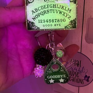 Puede incluir: Un carrete de insignia de tablero Ouija brillante con un dije de planchette. El tablero Ouija tiene el alfabeto, n&uacute;meros y las palabras "S&iacute;", "No" y "Adi&oacute;s". El dije de planchette tiene las palabras "S&iacute;", "No" y "Adi&oacute;s" con dos estrellas.