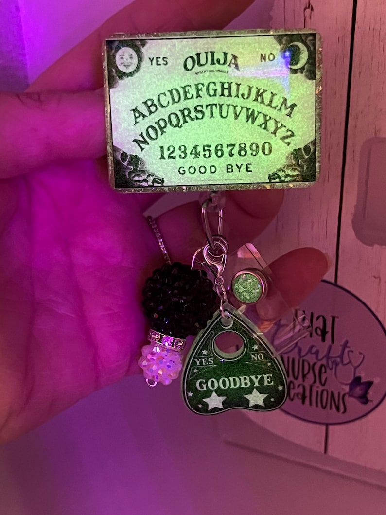 Puede incluir: Un carrete de insignia de tablero Ouija brillante con un dije de planchette. El tablero Ouija tiene el alfabeto, n&uacute;meros y las palabras "S&iacute;", "No" y "Adi&oacute;s". El dije de planchette tiene las palabras "S&iacute;", "No" y "Adi&oacute;s" con dos estrellas.