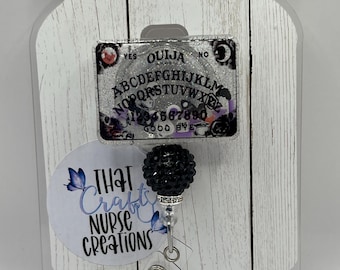 Puede incluir: Un fondo de madera blanco con un marco de plástico transparente. Dentro del marco hay una tabla Ouija en blanco y negro con un amuleto negro y plateado unido a un portagafete retráctil. El amuleto es una bola de diamantes de imitación negros con una mariposa plateada en el lateral. El portagafete está unido a un círculo blanco con el texto "That Crafty Nurse Creations".
