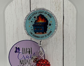 May include: A circular badge reel featuring a light blue background with a cartoon dumpster on fire and the text "THIS LITTLE LIGHT OF MINE I'M GONNA LET IT SHINE". Below the reel hangs a charm with a large red glitter bead and a smaller orange bead.