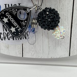 Puede incluir: Un carrete para placa con un dije de tablero Ouija en blanco y negro que dice "Goodbye". El dije est&aacute; unido a un carrete retr&aacute;ctil con un acento de cuentas negras y una cuenta iridiscente m&aacute;s peque&ntilde;a.