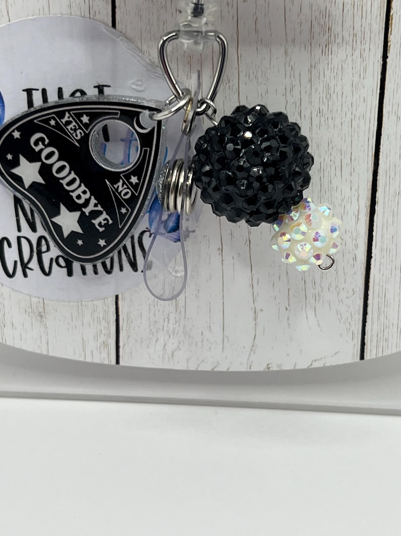 Puede incluir: Un carrete para placa con un dije de tablero Ouija en blanco y negro que dice "Goodbye". El dije est&aacute; unido a un carrete retr&aacute;ctil con un acento de cuentas negras y una cuenta iridiscente m&aacute;s peque&ntilde;a.