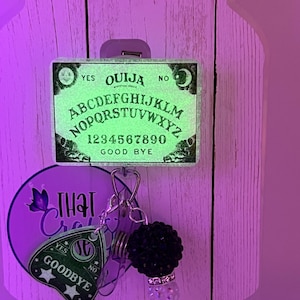 Puede incluir: Un carrete de insignia de tablero Ouija verde brillante con las palabras "S&Iacute;", "NO" y "ADI&Oacute;S". El carrete tambi&eacute;n presenta un dije con una bola de cuentas negras y un dije decorativo transparente. El fondo es una superficie de madera morada clara.