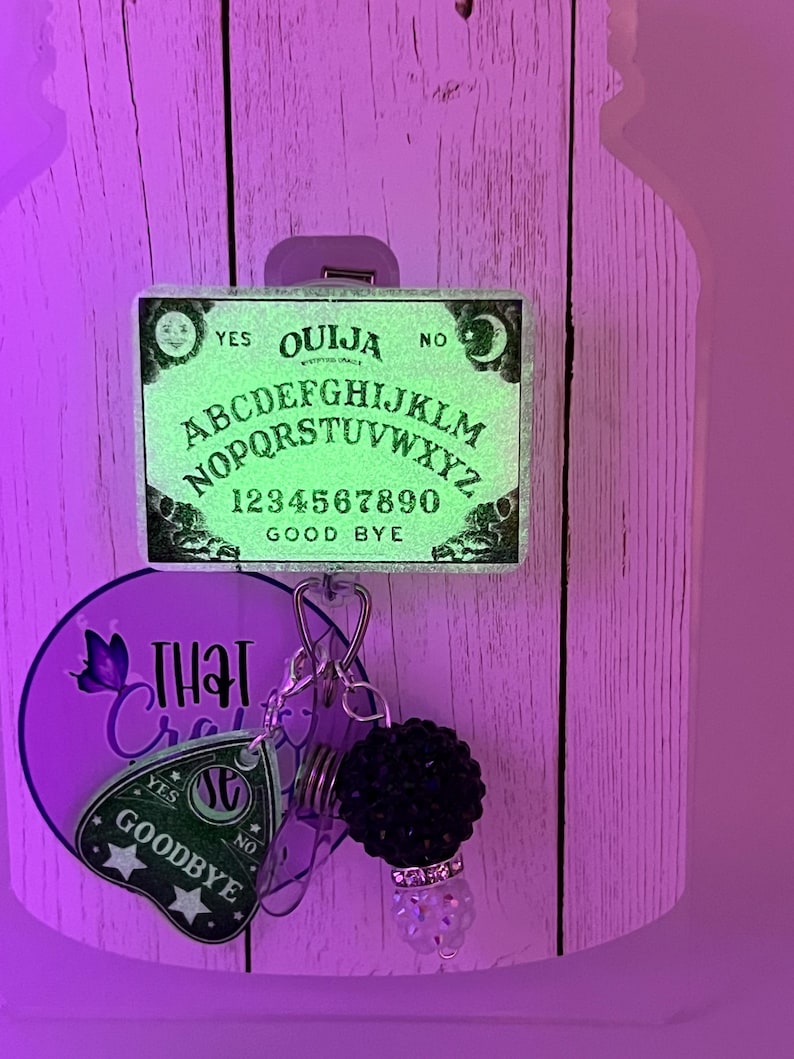 Puede incluir: Un carrete de insignia de tablero Ouija verde brillante con las palabras "S&Iacute;", "NO" y "ADI&Oacute;S". El carrete tambi&eacute;n presenta un dije con una bola de cuentas negras y un dije decorativo transparente. El fondo es una superficie de madera morada clara.