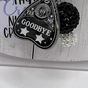 Puede incluir: Un dije de tablero Ouija negro y plateado con la palabra "GOODBYE" y detalles de estrellas. El dije est&aacute; unido a un llavero con una bola de cuentas negras y una cuenta blanca iridiscente.