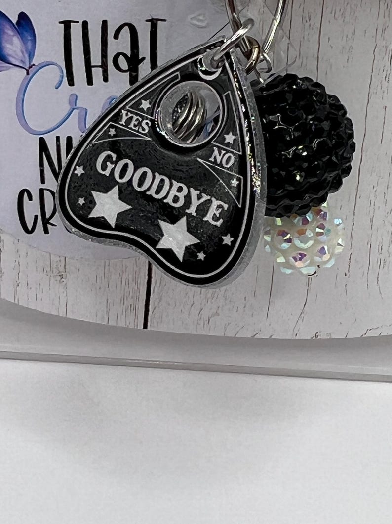 Puede incluir: Un dije de tablero Ouija negro y plateado con la palabra "GOODBYE" y detalles de estrellas. El dije est&aacute; unido a un llavero con una bola de cuentas negras y una cuenta blanca iridiscente.
