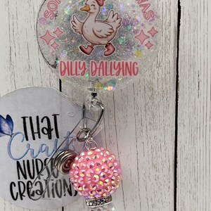 May include: A clear badge reel with a cartoon duck wearing a pink bow and shoes, with the text "Sorry I'm late I was dilly dallying". The reel has a pink and iridescent charm.