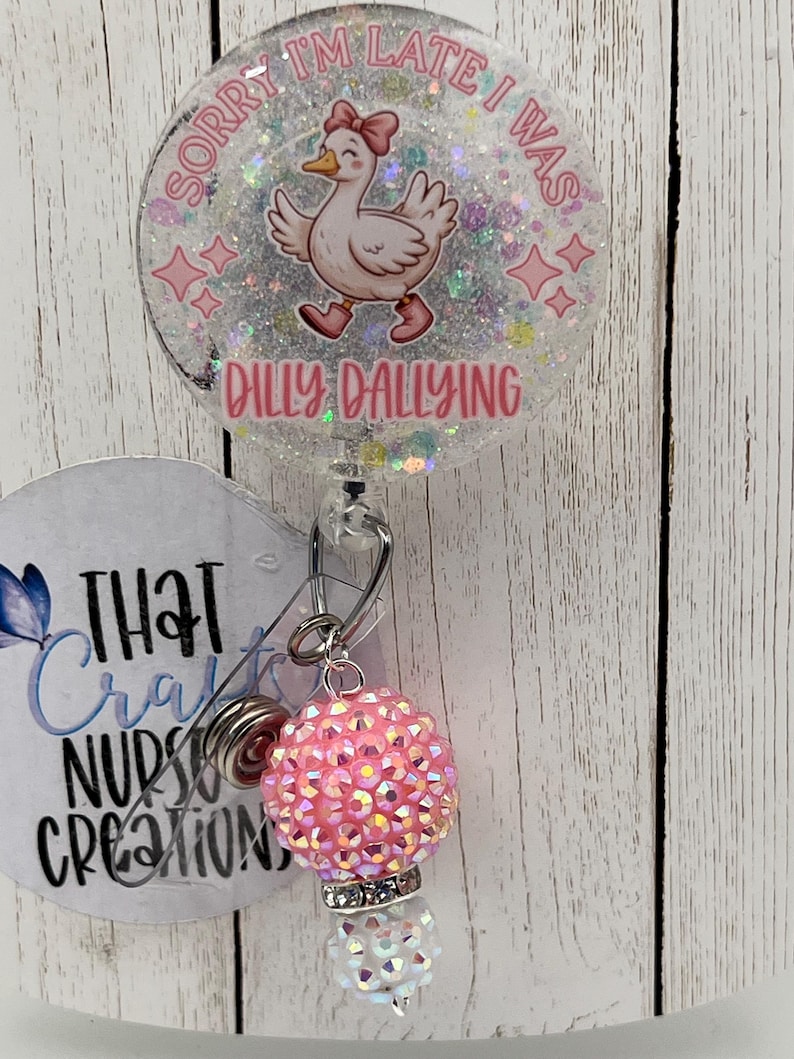 May include: A clear badge reel with a cartoon duck wearing a pink bow and shoes, with the text "Sorry I'm late I was dilly dallying". The reel has a pink and iridescent charm.