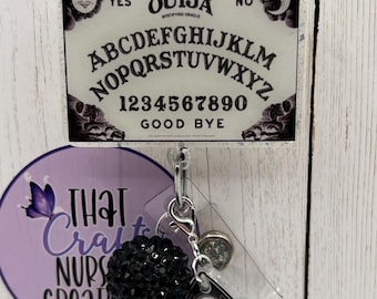 Puede incluir: Un carrete de insignia con un diseño de tablero Ouija, que muestra el alfabeto, los números y las palabras "Sí", "No" y "Adiós". Incluye un dije negro e iridiscente y un botón redondo con un diseño verde.