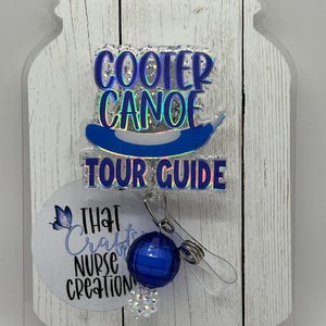 May include: A badge reel with the text "COOTER CANOE TOUR GUIDE" in blue and iridescent lettering. The reel features a blue canoe graphic and a round, blue bead with a silver clip. A white circle with the text "THAT Crafts NURSE CREATION" is also present.