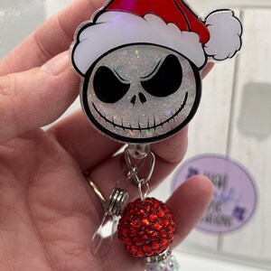May include: A badge reel featuring a glittery white skull character wearing a red and white Santa hat. Below the character, a metal clip is attached to a dangling charm consisting of a large red rhinestone bead and a smaller iridescent bead.