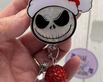 May include: A badge reel featuring a glittery white skull character wearing a red and white Santa hat. Below the character, a metal clip is attached to a dangling charm consisting of a large red rhinestone bead and a smaller iridescent bead.