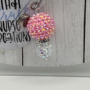 May include: A pink and iridescent badge reel with a rhinestone-covered ball and a smaller, white, iridescent ball. The reel has a silver-colored clip and a clear plastic backing. The text on the backing is partially visible.