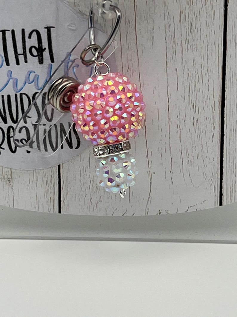 May include: A pink and iridescent badge reel with a rhinestone-covered ball and a smaller, white, iridescent ball. The reel has a silver-colored clip and a clear plastic backing. The text on the backing is partially visible.