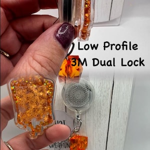 May include: Clear badge reel with orange glitter accents and the text "Low Profile 3M Dual Lock Interchangeable". The reel has a clear, rectangular shape with a decorative orange glitter design.