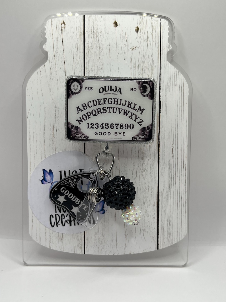 Puede incluir: Un portatarjetas con un dise&ntilde;o de tablero Ouija. La insignia rectangular presenta el alfabeto, los n&uacute;meros y las palabras "Yes", "No" y "Good Bye". Un dije con una planchette y detalles de cuentas negras cuelga debajo.
