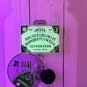 Puede incluir: Un adorno de pared decorativo con tem&aacute;tica de tablero Ouija. El tablero presenta el alfabeto, n&uacute;meros y las palabras &laquo;S&iacute;&raquo;, &laquo;No&raquo; y &laquo;Adi&oacute;s&raquo;. Un llavero con un dije de planchette y un accesorio de cuentas negras cuelga debajo. El fondo es un dise&ntilde;o de vetas de madera clara.