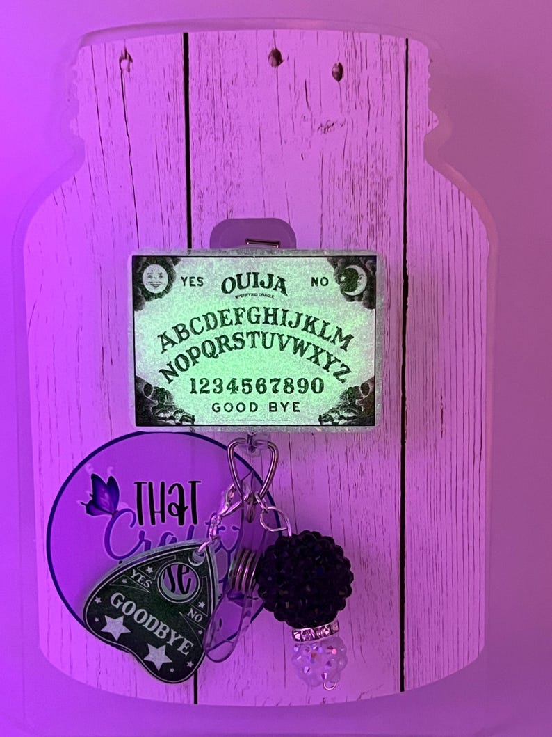 Puede incluir: Un adorno de pared decorativo con tem&aacute;tica de tablero Ouija. El tablero presenta el alfabeto, n&uacute;meros y las palabras &laquo;S&iacute;&raquo;, &laquo;No&raquo; y &laquo;Adi&oacute;s&raquo;. Un llavero con un dije de planchette y un accesorio de cuentas negras cuelga debajo. El fondo es un dise&ntilde;o de vetas de madera clara.
