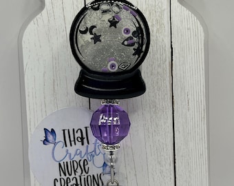 Puede incluir: Un carrete de insignia transparente en forma de tarro con un diseño de globo de nieve negro y plateado. El globo de nieve presenta estrellas, lunas y otras formas. El carrete tiene una cuenta púrpura y el texto "THAT Craft NURSE CREATIONS".