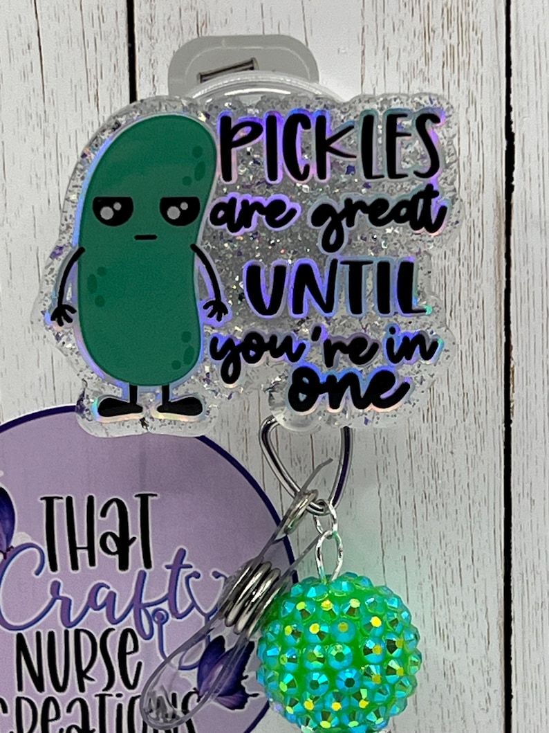 May include: A badge reel with a green pickle cartoon and the text "PICKLES are great UNTIL you're in one." The badge reel has a clear plastic clip and a green and blue rhinestone charm.