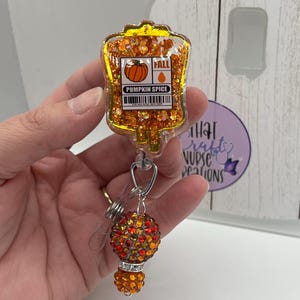 May include: A badge reel featuring a clear, glitter-filled resin IV bag shape with an orange pumpkin graphic and the text PUMPKIN SPICE and FALL. Below the bag, a silver metal clasp connects to two round beads covered in orange and red rhinestones.