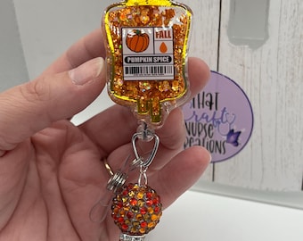 May include: A badge reel featuring a clear, glitter-filled resin IV bag shape with an orange pumpkin graphic and the text PUMPKIN SPICE and FALL. Below the bag, a silver metal clasp connects to two round beads covered in orange and red rhinestones.