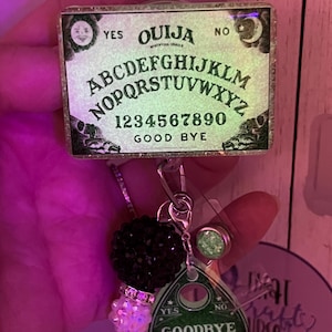 Puede incluir: Un portatarjetas con un dise&ntilde;o de tablero Ouija en miniatura, que incluye el alfabeto, n&uacute;meros y las palabras "YES", "NO" y "GOOD BYE". Incluye un dije de cuentas negras, un dije de planchette y un acento de purpurina verde.