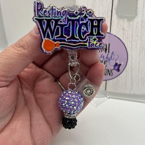 May include: A badge reel featuring a purple glittery topper with the text Resting Witch Face in black and holographic lettering, accented by an orange broomstick icon. A dangling charm consists of a light purple and a black rhinestone bead.