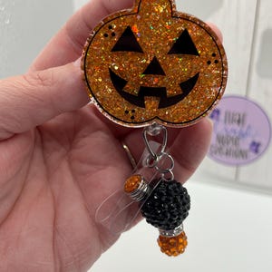 May include: A badge reel featuring a circular orange pumpkin face with black triangular eyes, nose, and a jagged mouth. The pumpkin is covered in orange glitter. Attached below are two small, round, textured beads in black and orange.
