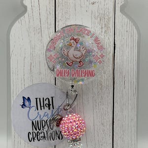 May include: A badge reel with a cartoon duck wearing a bow and the text "Sorry I'm late I was dilly dallying." The reel has a pink and white beaded charm and a smaller tag with the text "That Craft Nurse Creations."