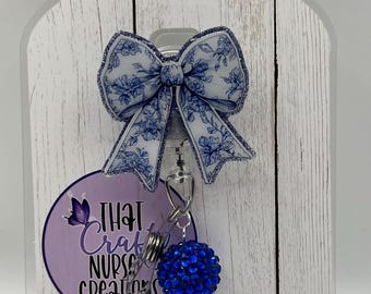 May include: A retractable badge reel with a blue and white floral bow, a blue rhinestone ball, and a clear, sparkling charm. The reel has the text "That Craft Nurse Creations" on a purple background.
