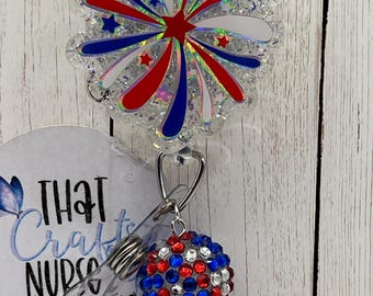 May include: A badge reel featuring a clear, glittery, scalloped-edge topper with a red, white, and blue firework design. Attached is a charm made of red, white, and blue rhinestones. The text "THAT CRAFTY NURSE CREATIONS" is visible on the backing.