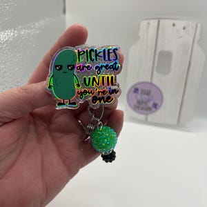 May include: A holographic badge reel with a green pickle cartoon and the text "Pickles are great until you're in one." The reel has a green, sparkly ball charm and a black base.