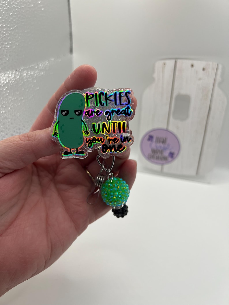 May include: A holographic badge reel with a green pickle cartoon and the text "Pickles are great until you're in one." The reel has a green, sparkly ball charm and a black base.