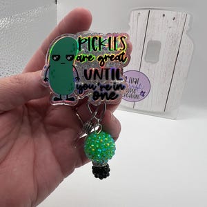 May include: A badge reel with a green pickle character and the text "Pickles are great until you're in one." The reel has a green and black beaded charm. The background is white.
