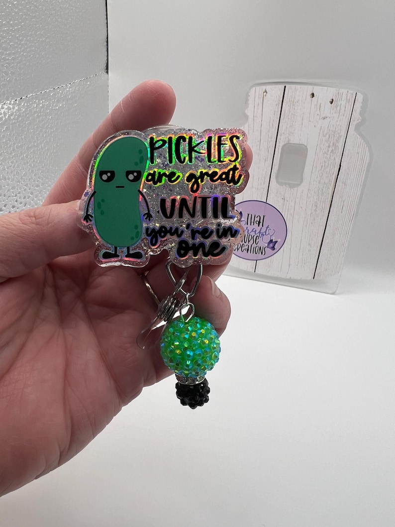 May include: A badge reel with a green pickle character and the text "Pickles are great until you're in one." The reel has a green and black beaded charm. The background is white.