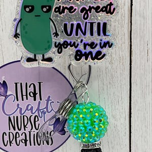 May include: A badge reel with a green pickle cartoon that says "Pickles are great until you're in one." The reel has a green and blue rhinestone bead and a black bead at the bottom. The reel also has the text "That Crafty Nurse Creations."