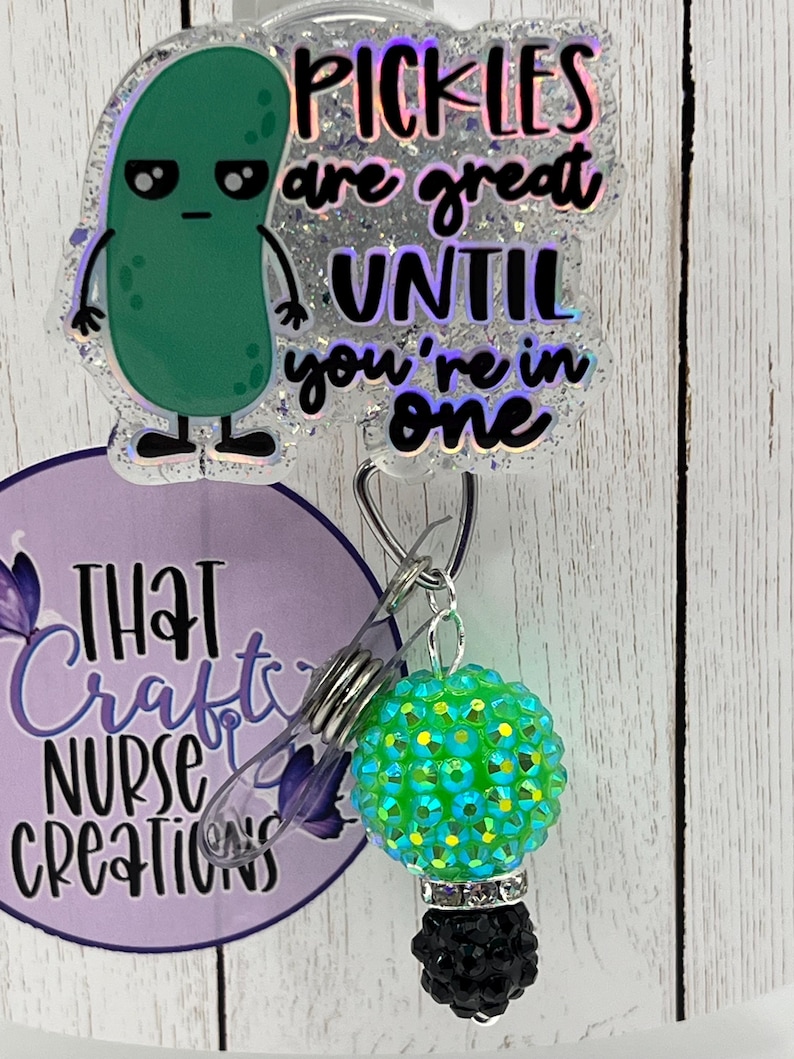 May include: A badge reel with a green pickle cartoon that says "Pickles are great until you're in one." The reel has a green and blue rhinestone bead and a black bead at the bottom. The reel also has the text "That Crafty Nurse Creations."