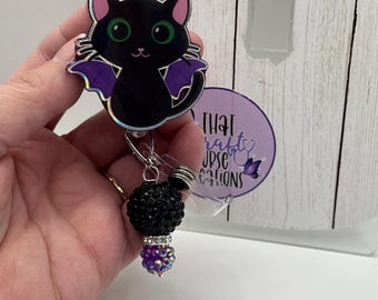 May include: A black cat badge reel with purple bat wings, green eyes, and pink inner ears. The accessory features a dangling charm with a large black rhinestone bead, a silver spacer, and a smaller purple rhinestone bead at the bottom.
