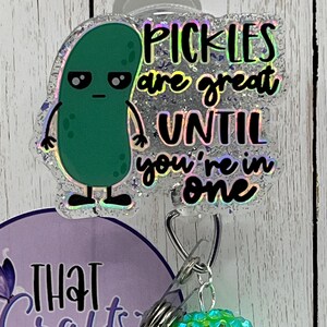 May include: A badge reel with a green pickle cartoon and the text "Pickles are great UNTIL you're in one." The reel also features a purple and white design with the text "THAT Crafty NURSE" and a green rhinestone charm.