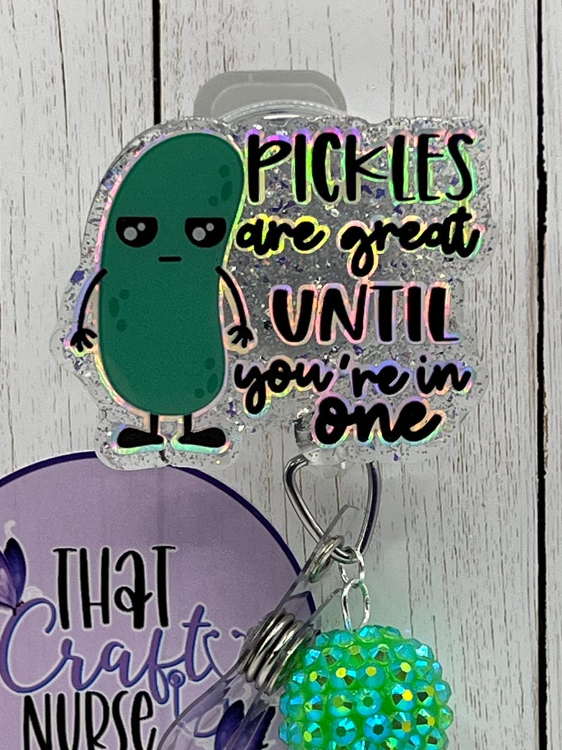 May include: A badge reel with a green pickle cartoon and the text "Pickles are great UNTIL you're in one." The reel also features a purple and white design with the text "THAT Crafty NURSE" and a green rhinestone charm.
