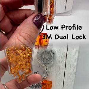 May include: Clear badge reel with orange glitter accents and the text "Low Profile 3M Dual Lock Interchangeable". The reel has a retractable cord and a decorative charm.