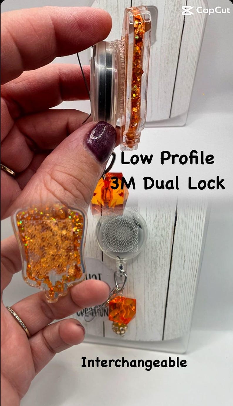 May include: Clear badge reel with orange glitter accents and the text "Low Profile 3M Dual Lock Interchangeable". The reel has a retractable cord and a decorative charm.
