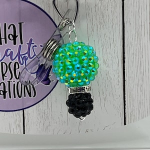 May include: A badge reel with a green and blue jeweled lightbulb charm and a black base. The badge reel has the text "you're the one" and "What a Crafty Nurse Creates" on a purple background.