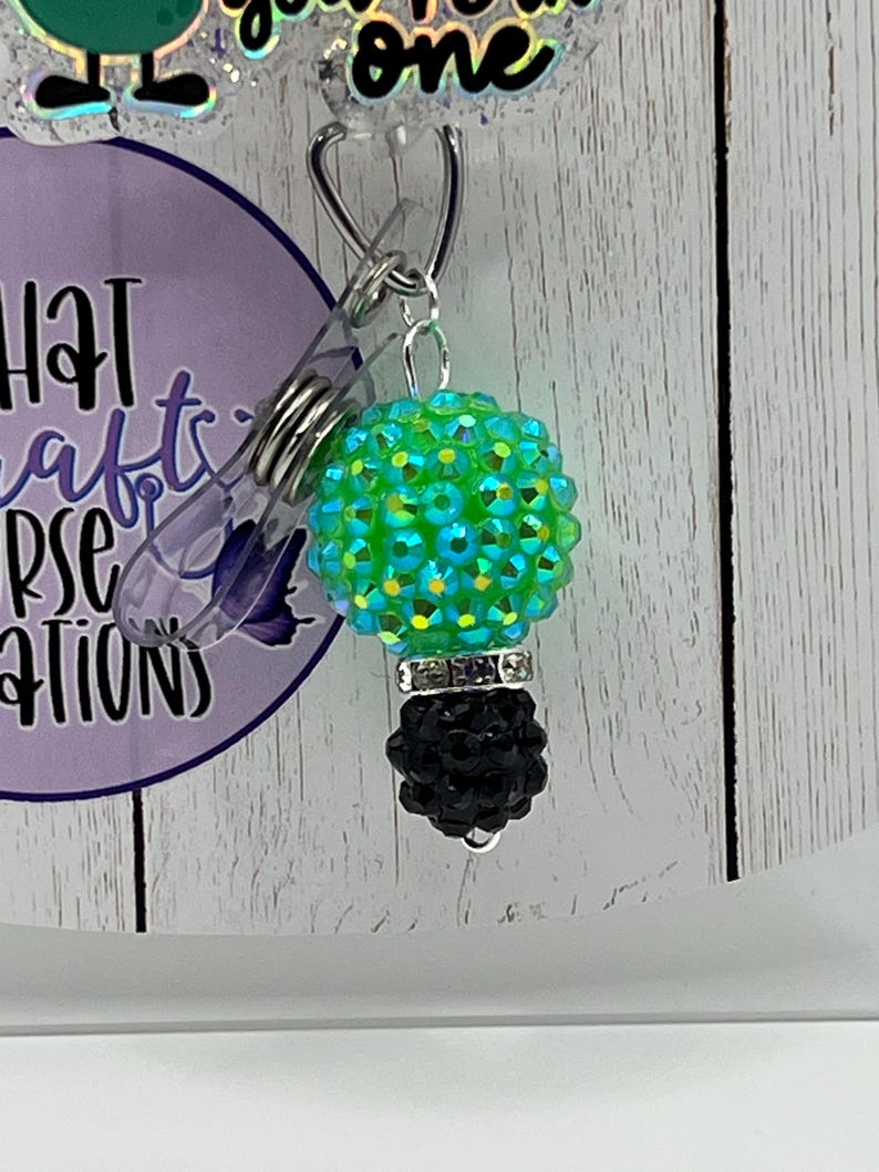 May include: A badge reel with a green and blue jeweled lightbulb charm and a black base. The badge reel has the text "you're the one" and "What a Crafty Nurse Creates" on a purple background.